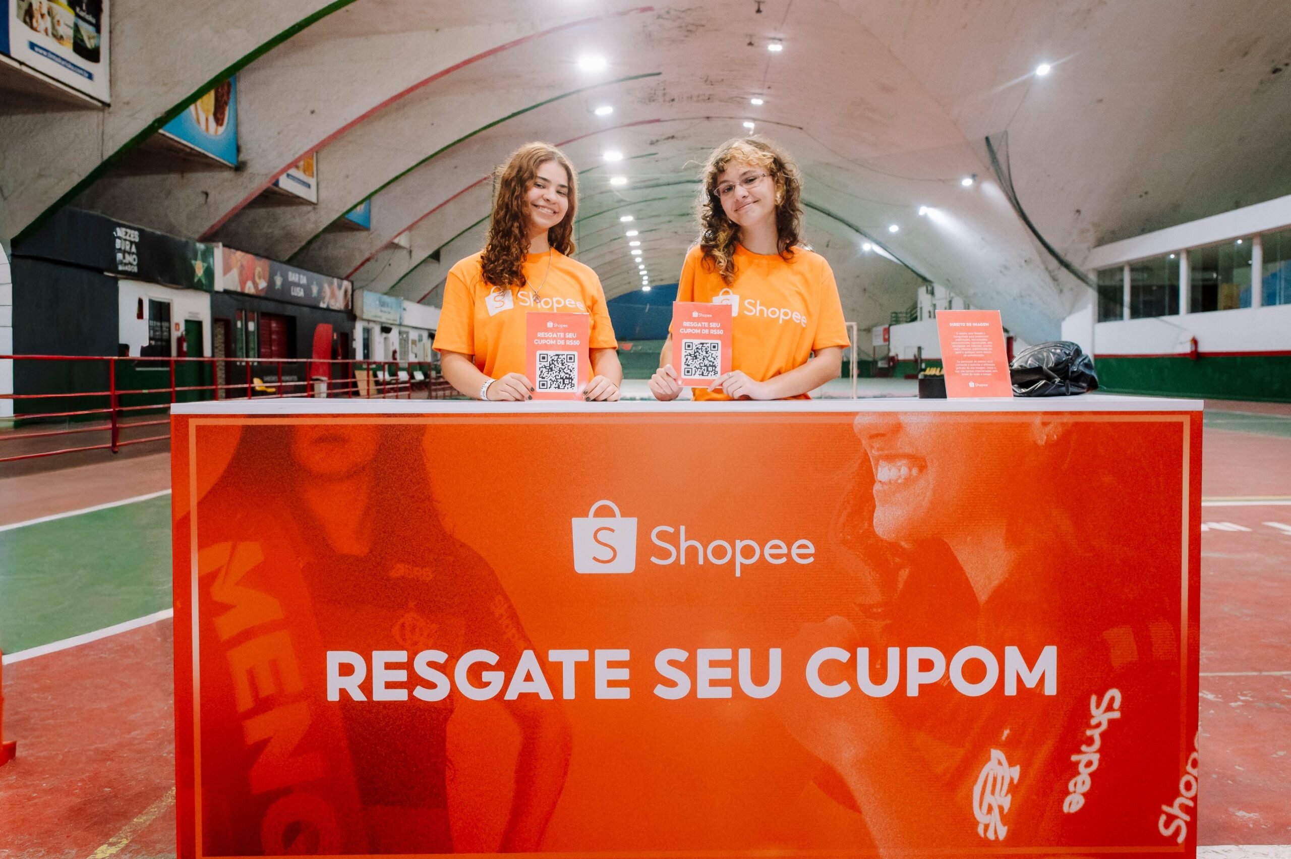 Balcão da Shopee no Estádio Luso-Brasileiro, com duas promotoras mostrando as placas de QR code para resgate do cupom de R$ 50