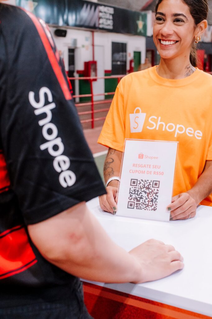 Promotora da Shopee no balcão da ativação mostra a placa com QR code para resgate do cupom de R$ 50 a um torcedor com a camisa do Flamengo, no Estádio Luso-Brasileiro